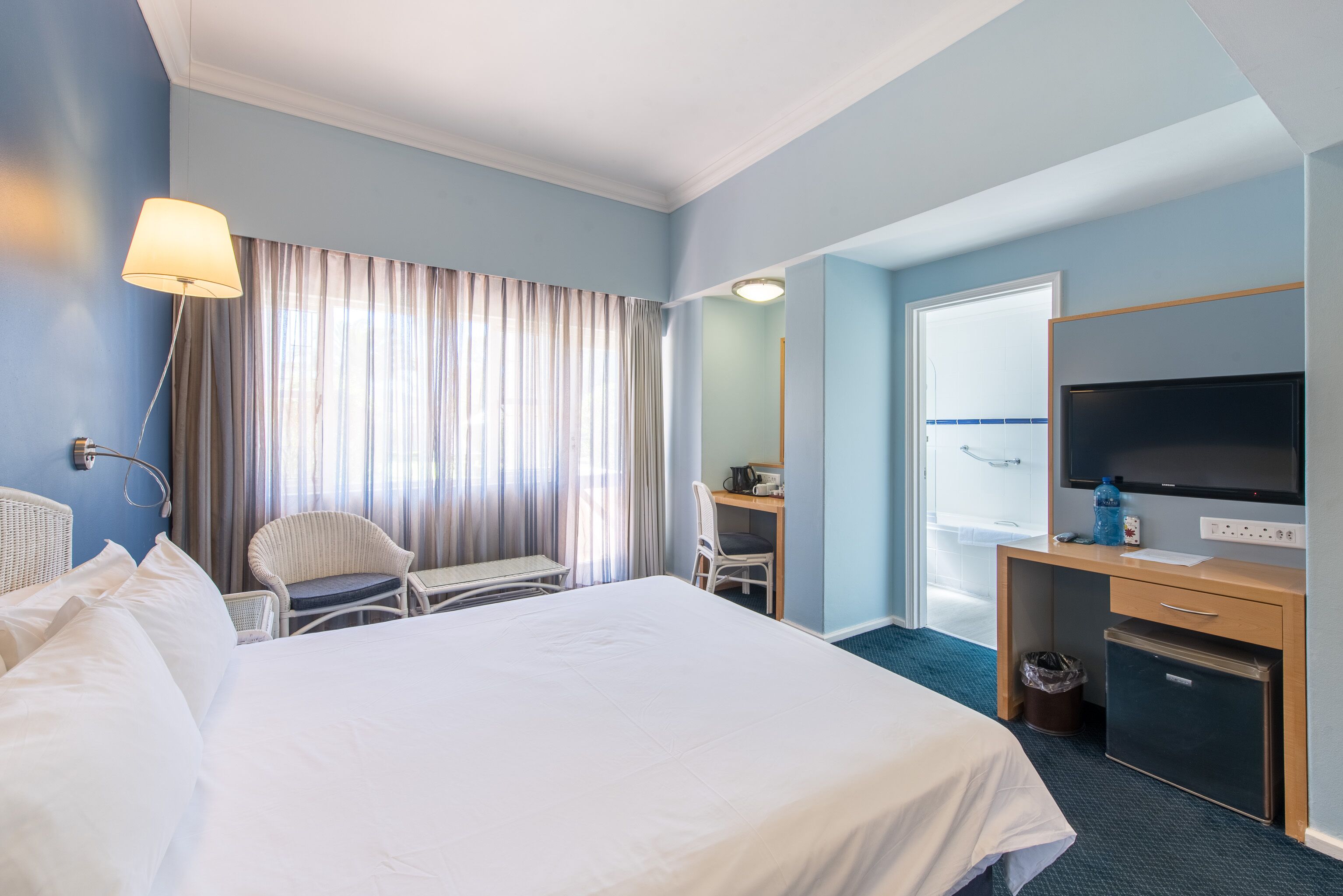 Habitación de hotel con una cama grande, un escritorio con silla, un televisor y un refrigerador pequeño.
