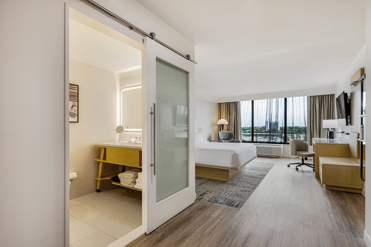 A hotel room with a bed, desk, chair, and a view of the city.