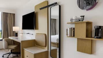 Une chambre d’hôtel équipée d’un bureau, d’une chaise et d’un grand miroir. On y trouve également un téléviseur fixé au mur, une machine à café et une étagère avec des articles de toilette.