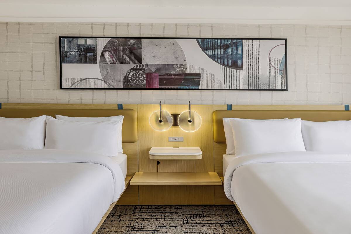 A hotel room with two beds, a bedside table, a wall-mounted artwork, and a light fixture.