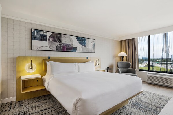 A hotel room with a large bed, a nightstand with a lamp, a chair, a large window with a view, and a wall-mounted artwork.
