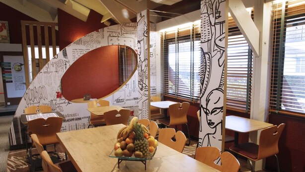A dining area with a wooden table, chairs, and a fruit bowl. There are walls with artistic designs and a window with blinds. - ibis Montbeliard (Montbeliard)