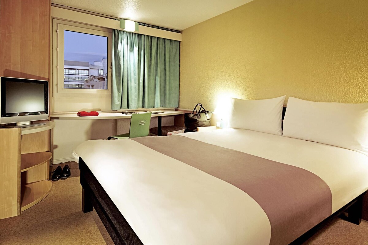 Una habitación de hotel con cama, escritorio, televisor y ventana con cortinas.