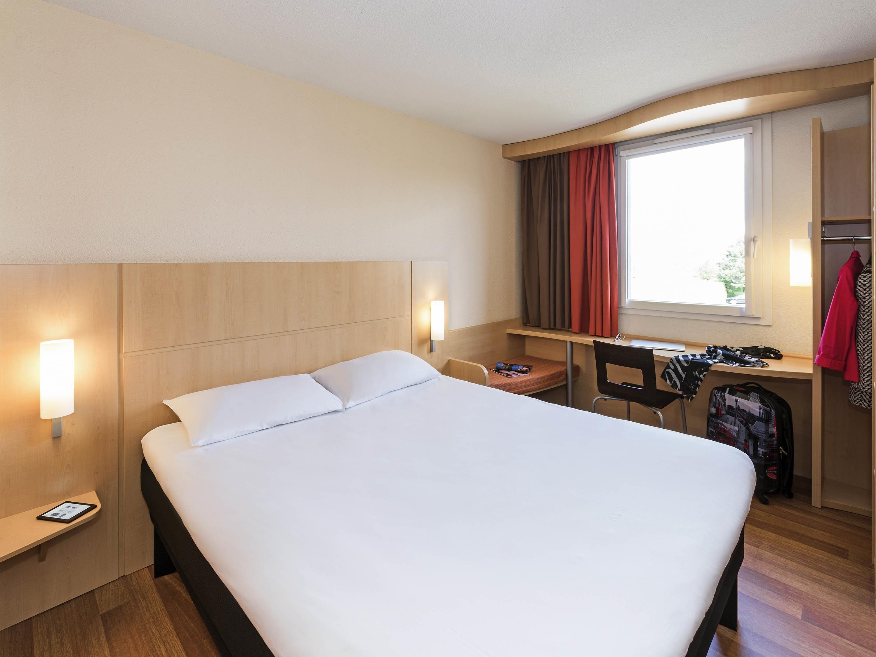Une chambre d’hôtel équipée d’un lit, d’un bureau, d’une chaise, d’une armoire et d’une fenêtre avec des rideaux.