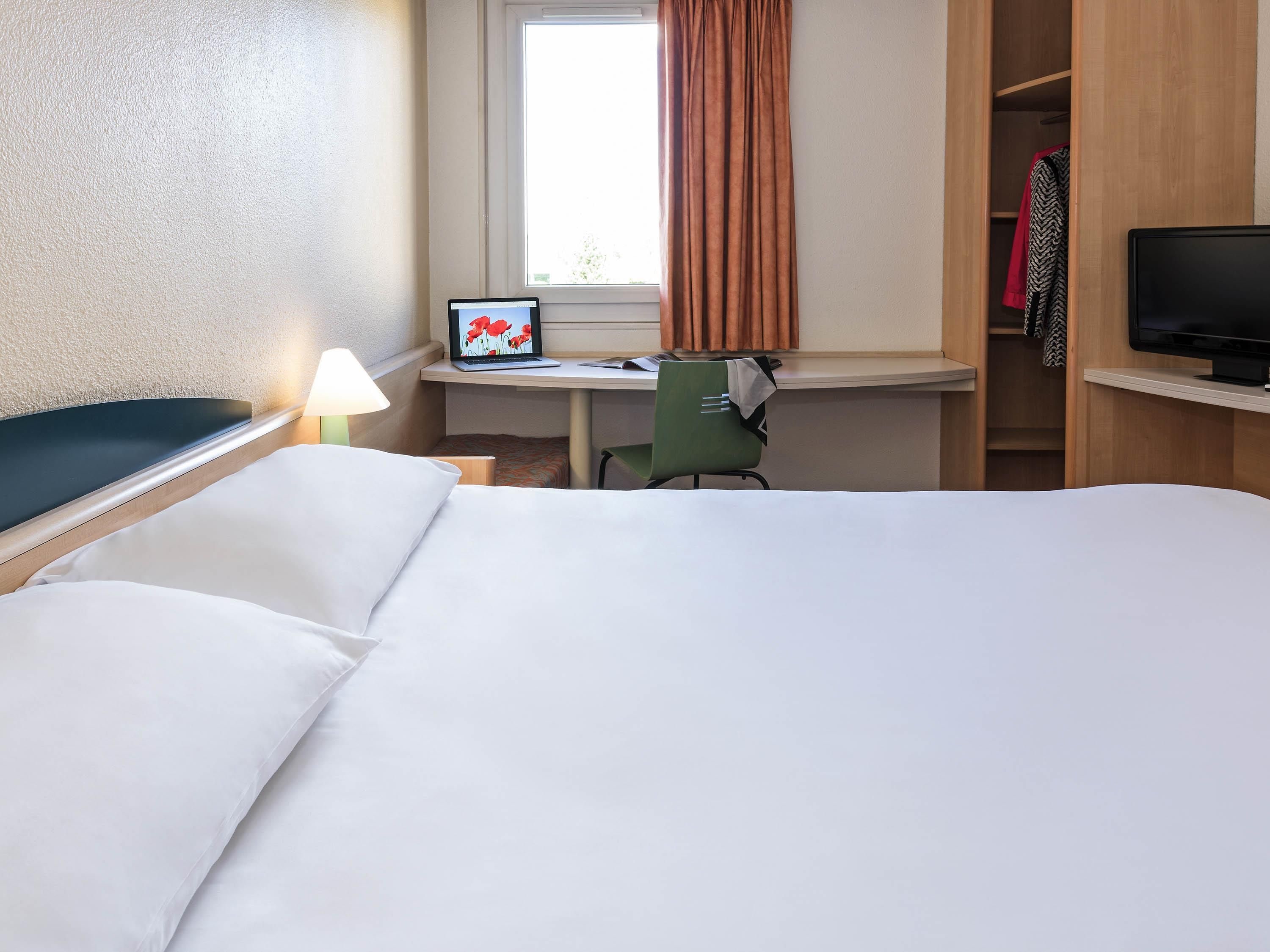 A hotel room with a bed, a desk with a laptop, a chair, a television, and a wardrobe.