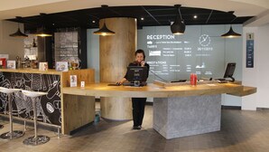 A reception desk with a large screen displaying reception information, a bar area with stools, and a modern interior design. - ibis Montbeliard (Montbeliard)