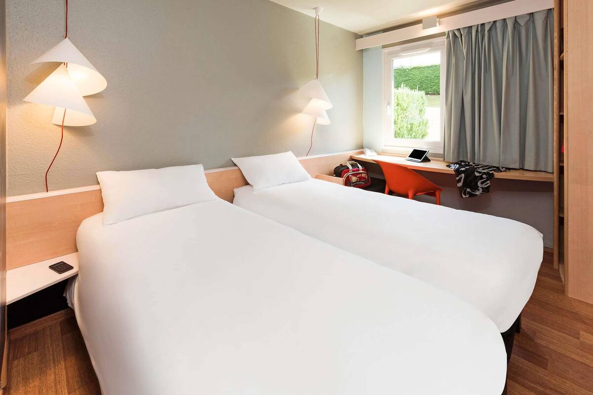 A hotel room with two beds, a desk, a chair, a window with curtains, and a lamp.