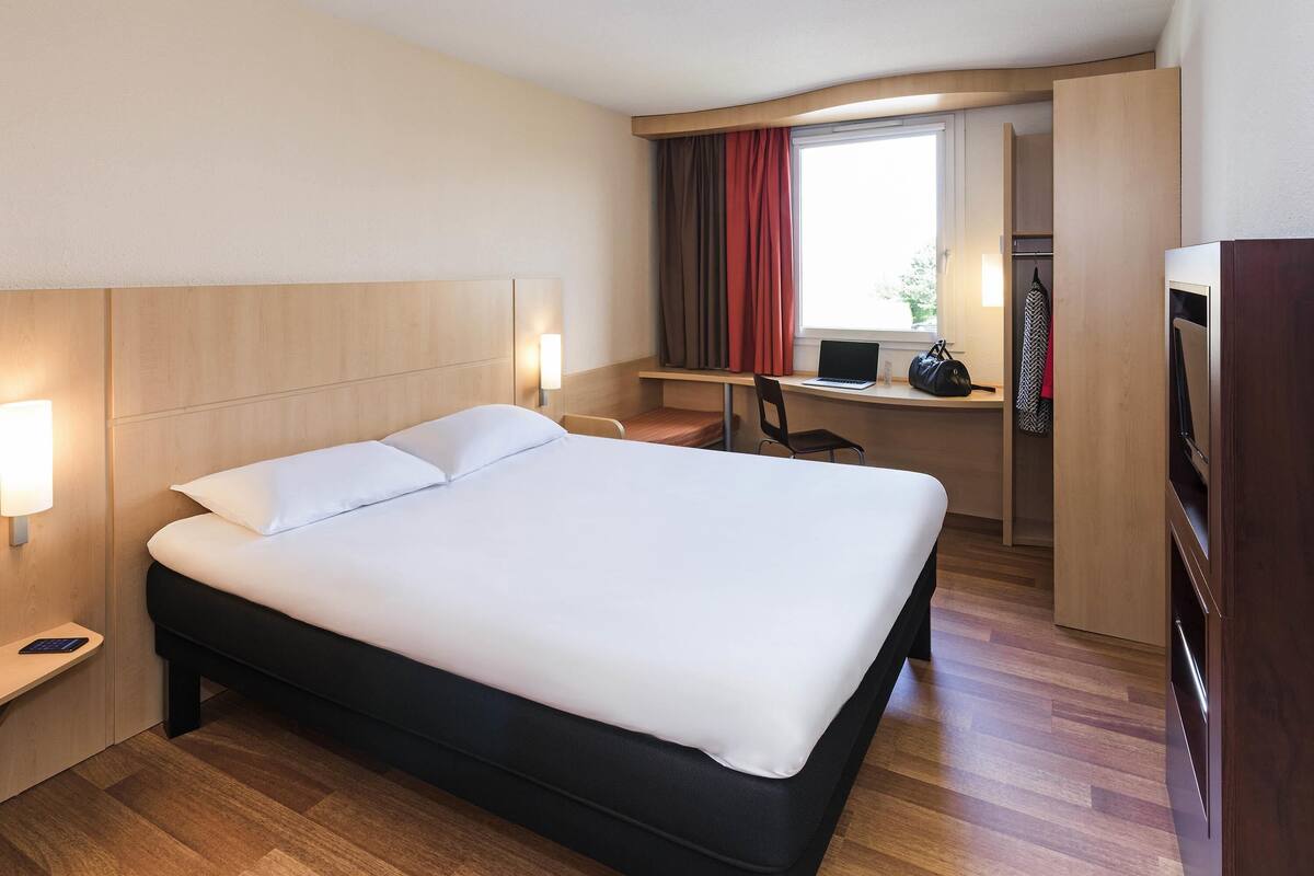 A hotel room with a large bed, a desk with a laptop, a chair, and a wardrobe.