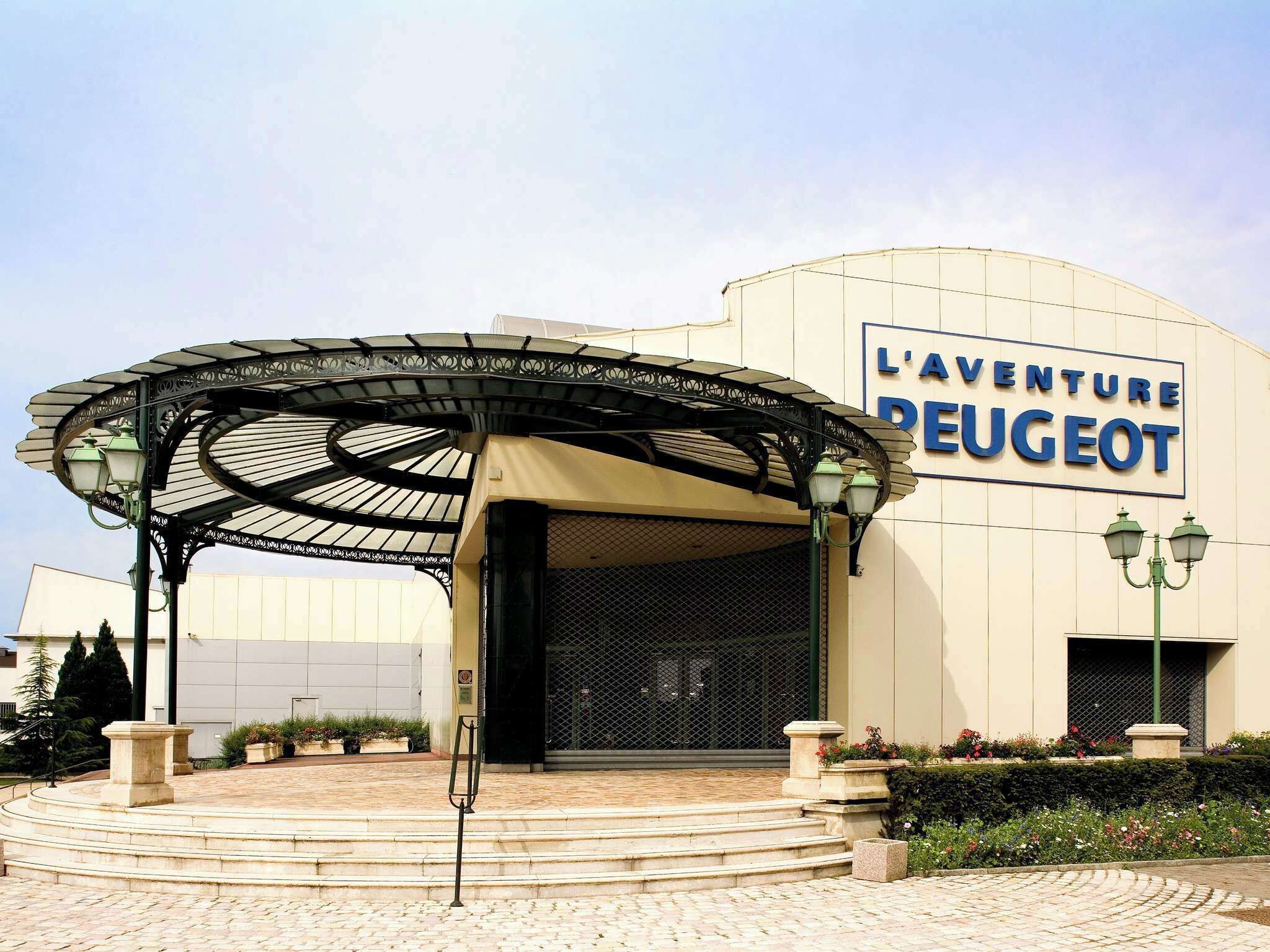 Ein Gebäude mit einem Schild, auf dem „L'Aventure Peugeot“ steht, und einem überdachten Eingangsbereich mit Stufen.