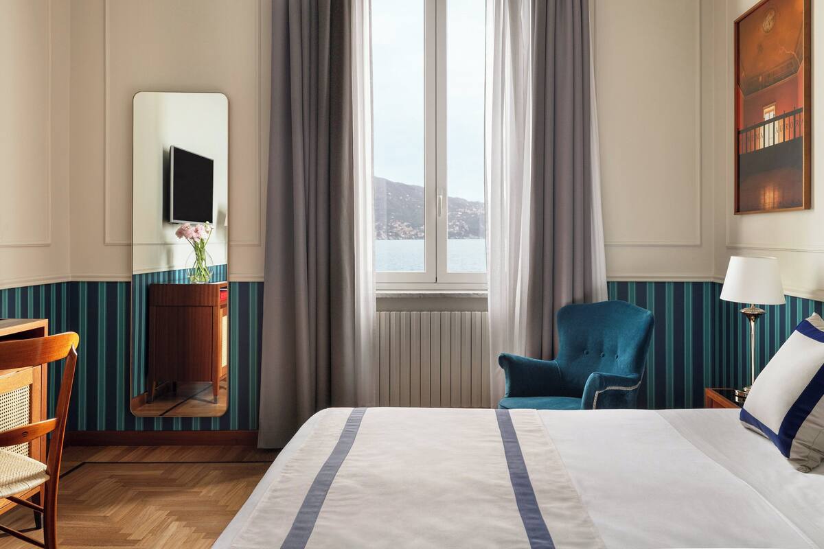 Hotellihuone, jossa on sänky, tuoli, työpöytä, peili, ikkuna verhoilla ja taulu seinällä.