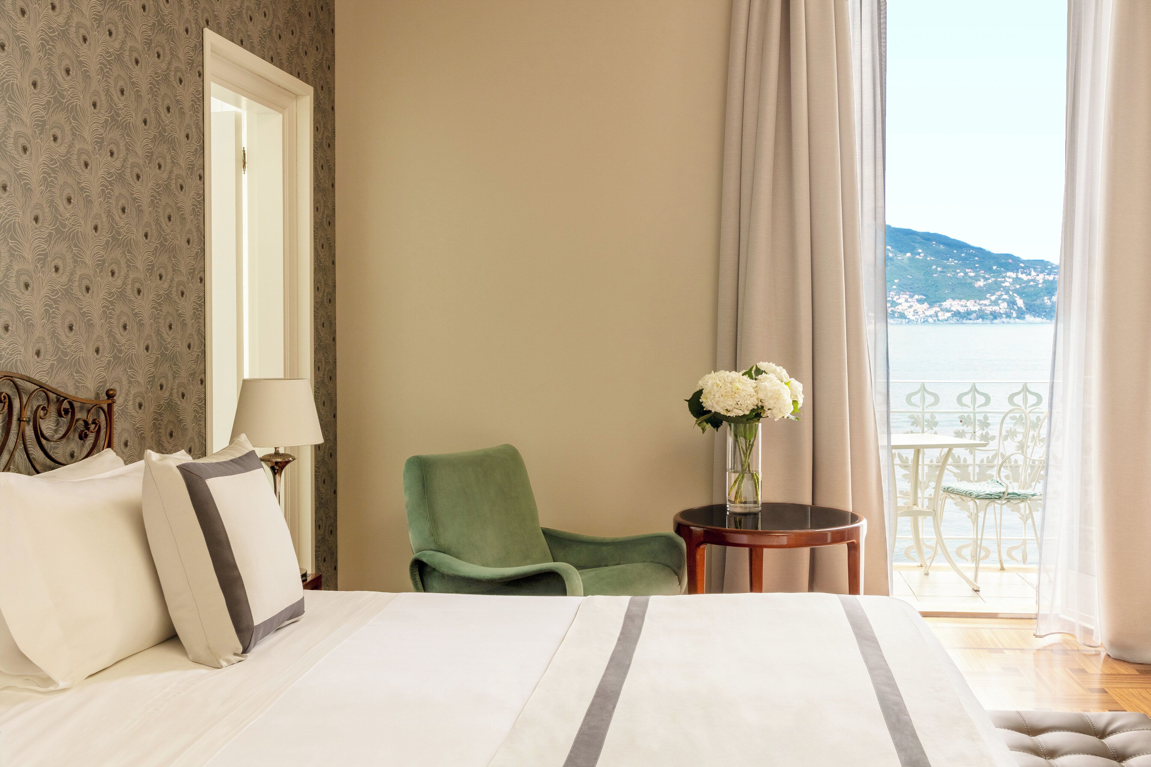 Une chambre d’hôtel avec un lit, une chaise, une petite table ornée d’un vase de fleurs et une vue sur la mer depuis un balcon.