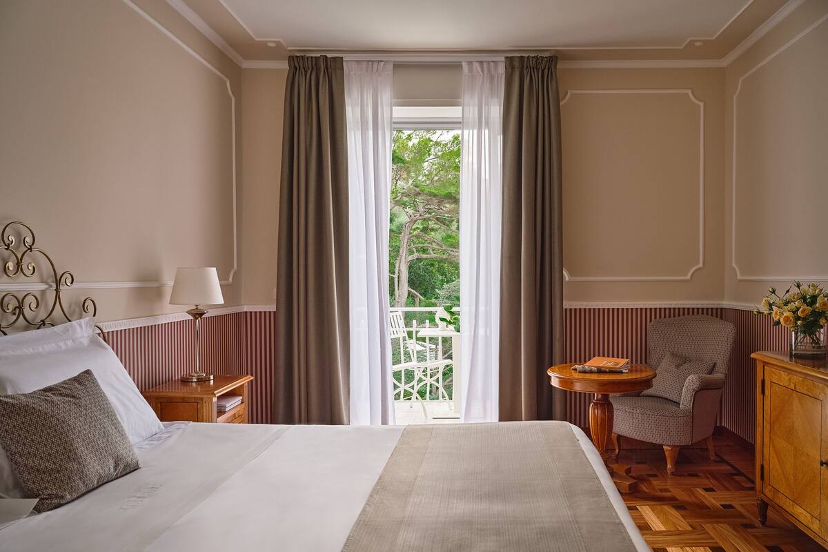 Hotellihuone, jossa on sänky, tuoli, pieni pöytä, yöpöytä ja näkymä ulos.
