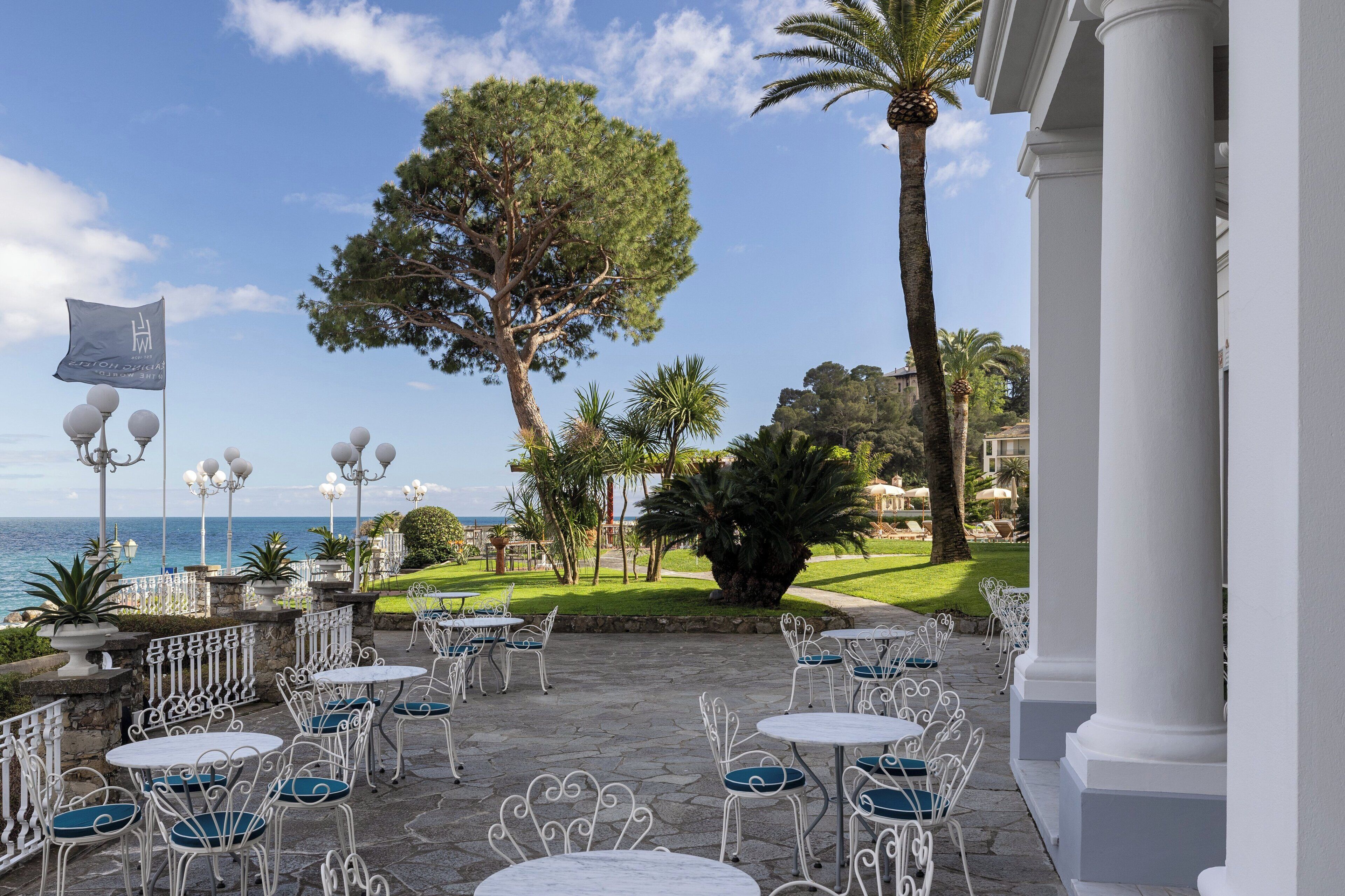 An outdoor patio with white metal chairs and tables, a flag, and a view of the sea.