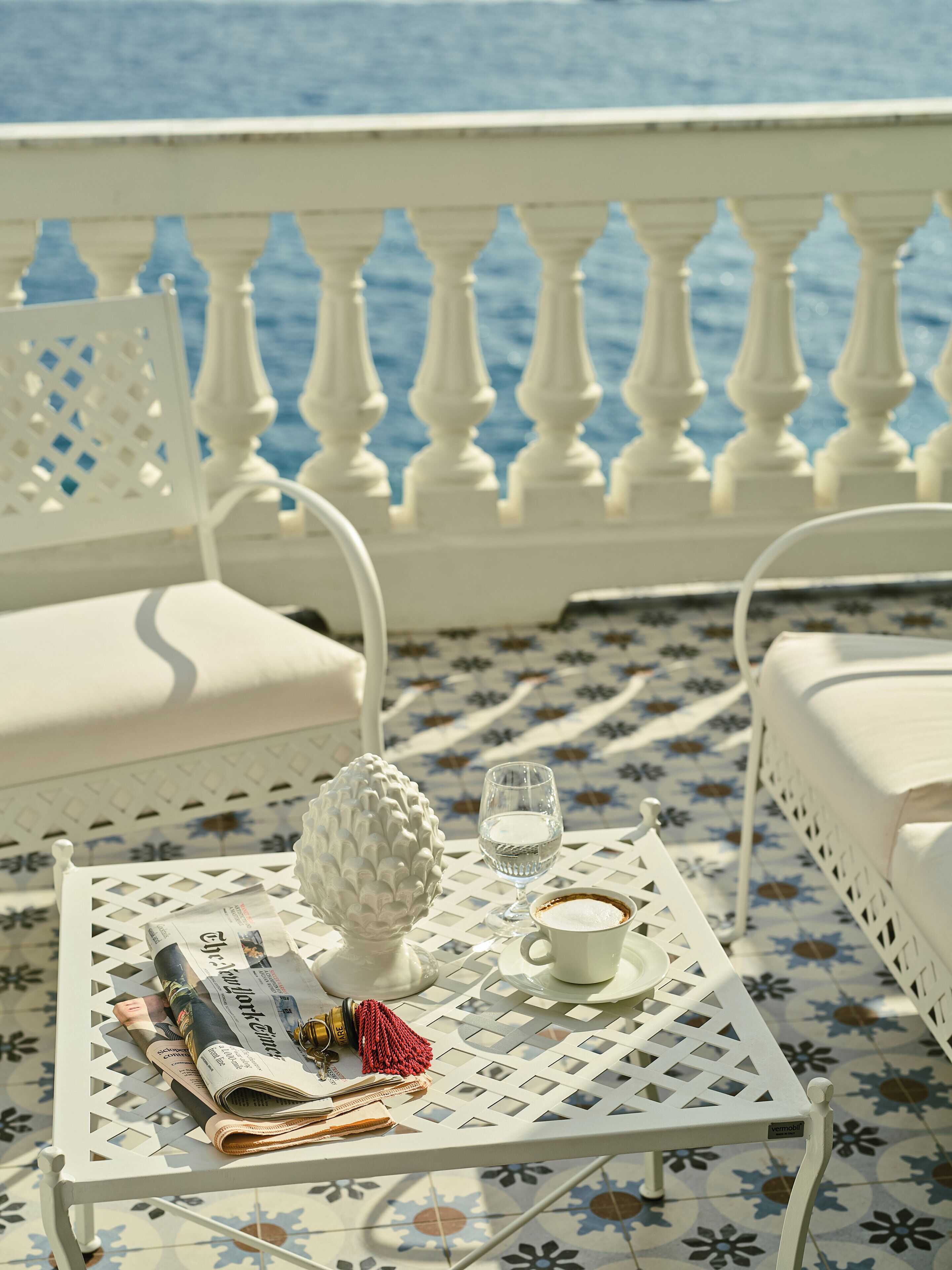 A white metal table with a newspaper, a glass of water, a cup of coffee, and a decorative item on a tiled floor with a view of the sea.