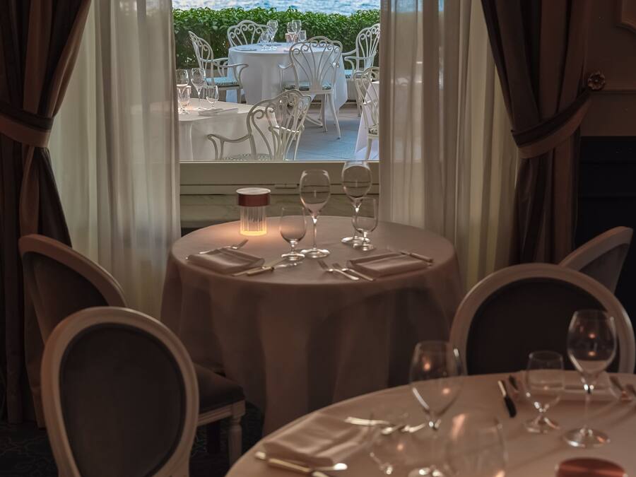 A dining table set for four with white chairs, a candle, and a view of the sea through large windows.