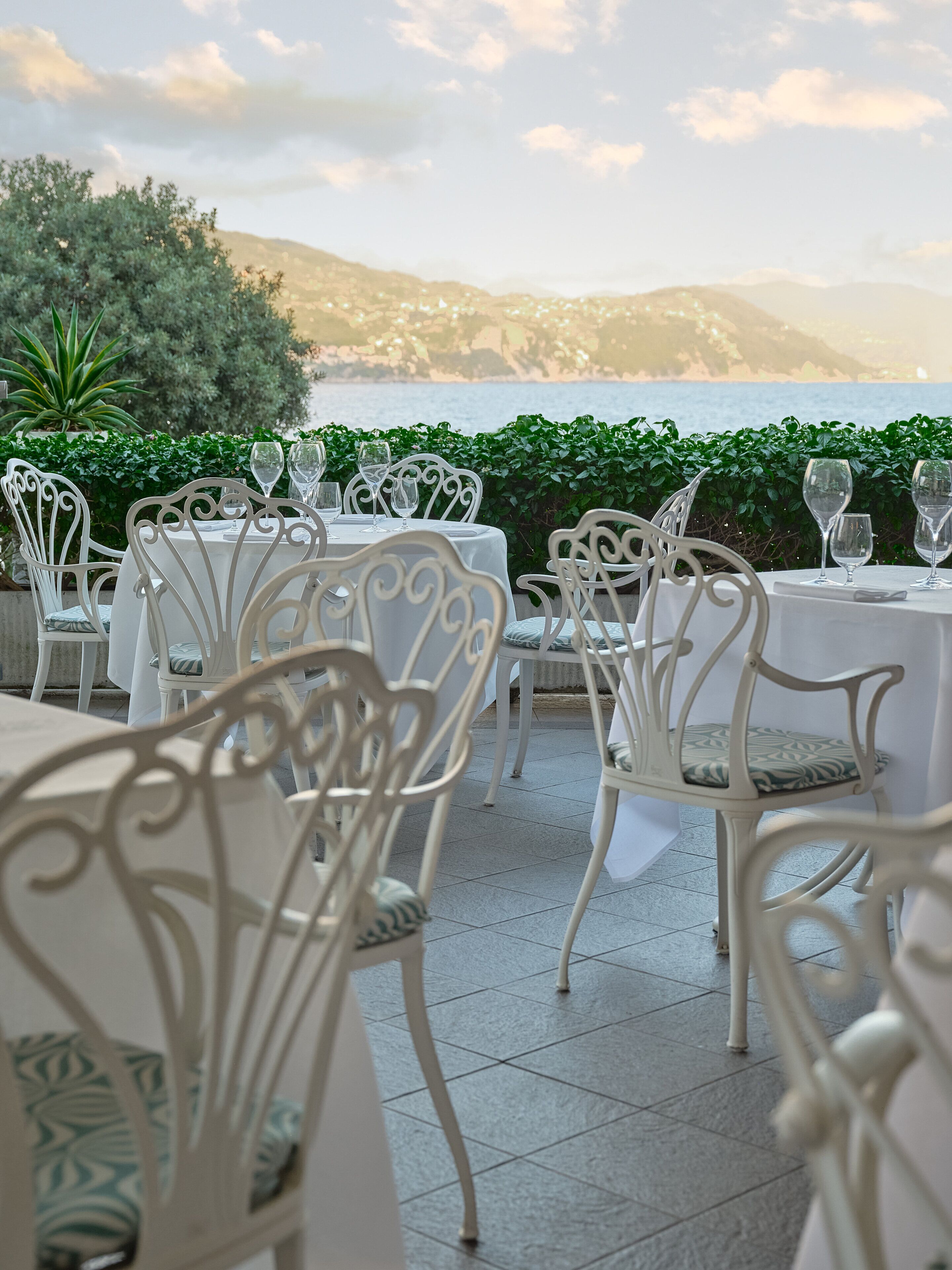 Un espace de restauration en plein air, avec des chaises et des tables en métal blanc, offrant une vue sur une étendue d’eau et des collines.