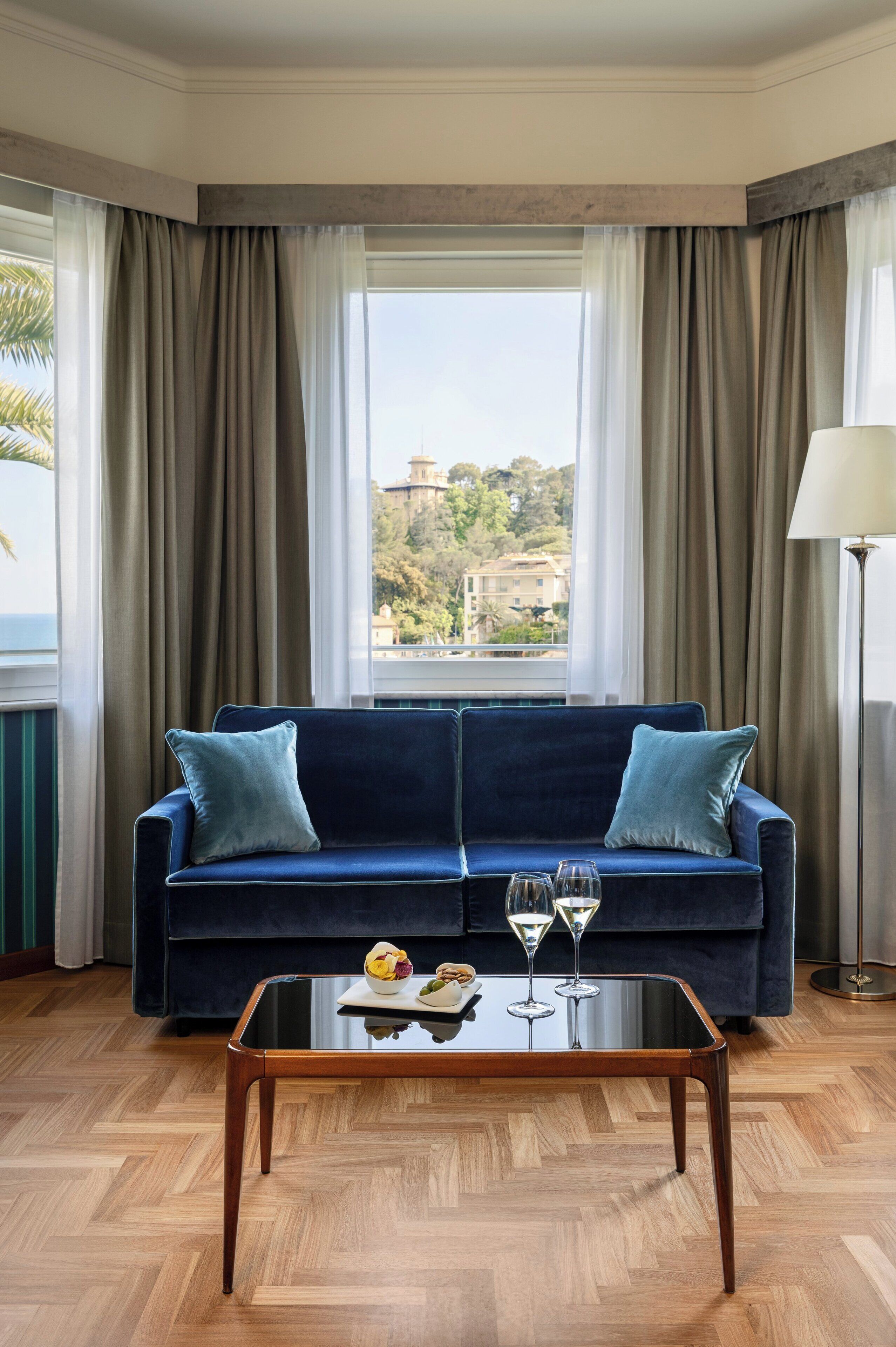 Ein Zimmer mit einem dunkelblauen Sofa, zwei Kissen, einem Glastisch mit einer Schale und zwei Weingläsern sowie Blick auf einen Hügel und Gebäude durch große Fenster mit beige farbenen Vorhängen.