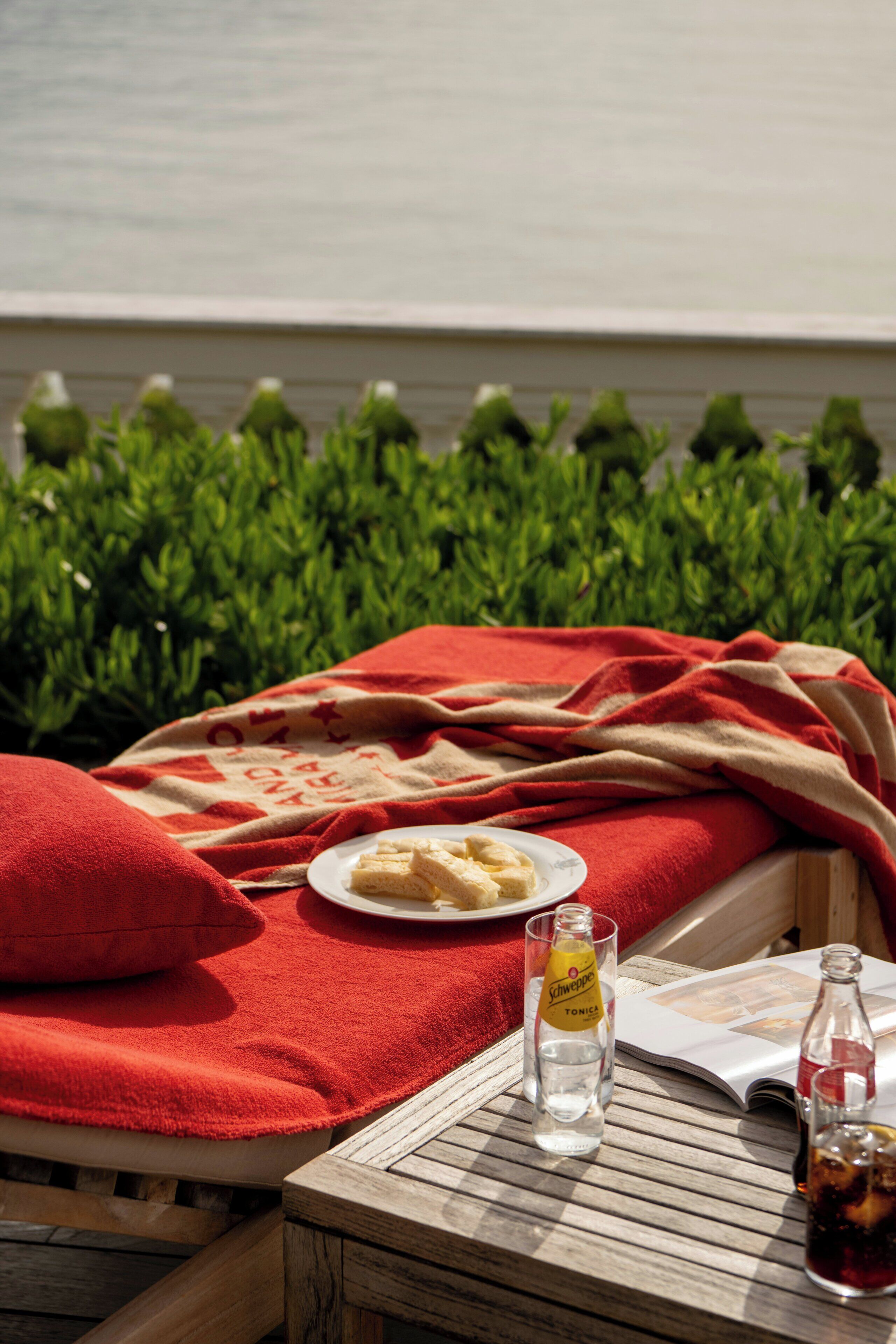 A sunlit outdoor seating area with a red blanket, a plate of food, a bottle of Schweppes tonic water, and a glass of water.