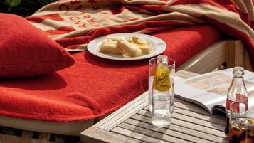 A sunlit outdoor seating area with a red blanket, a plate of food, a bottle of Schweppes tonic water, and a glass of water.