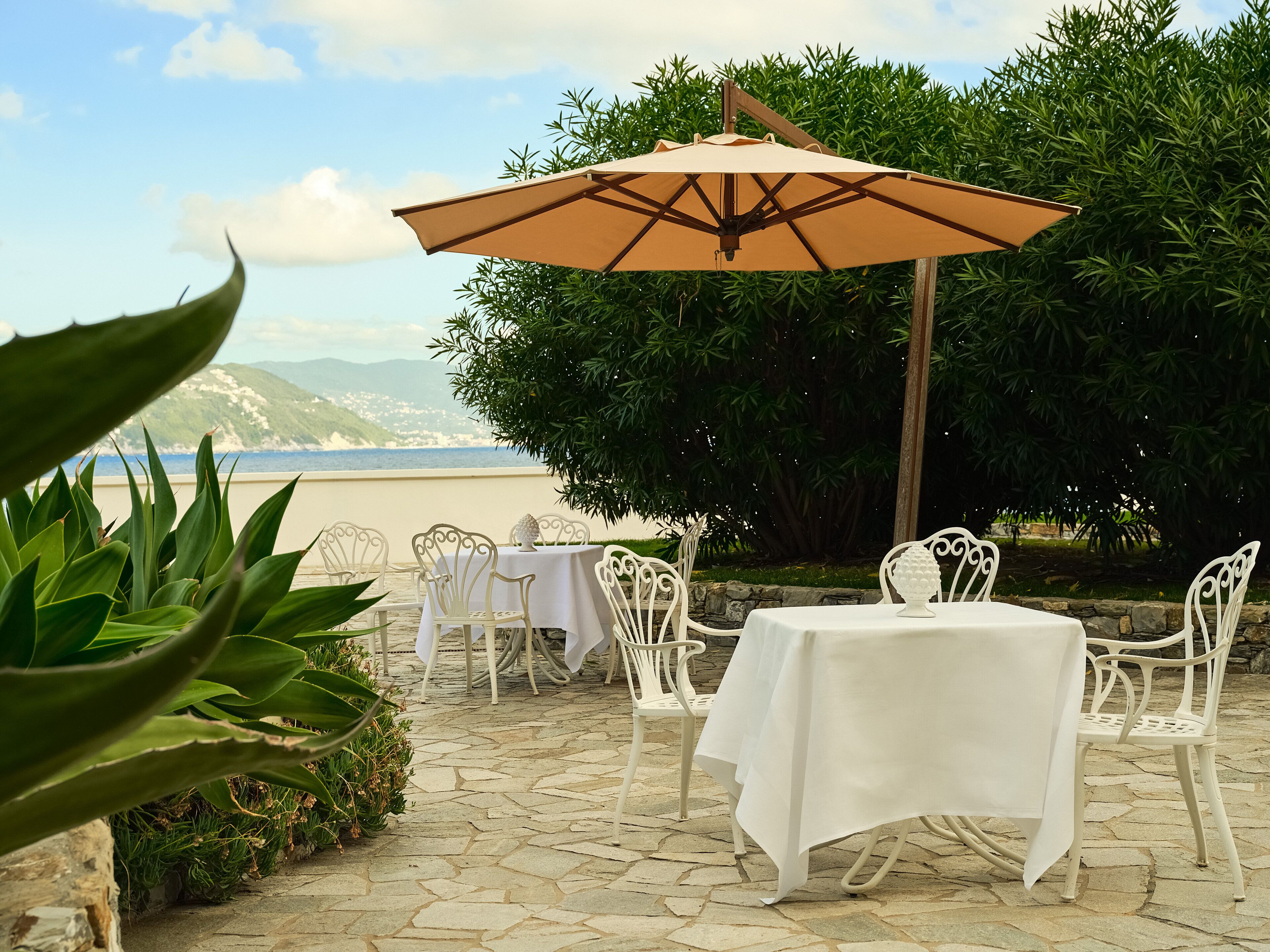 A patio with white metal chairs and tables, a beige umbrella, and a view of the sea and mountains.