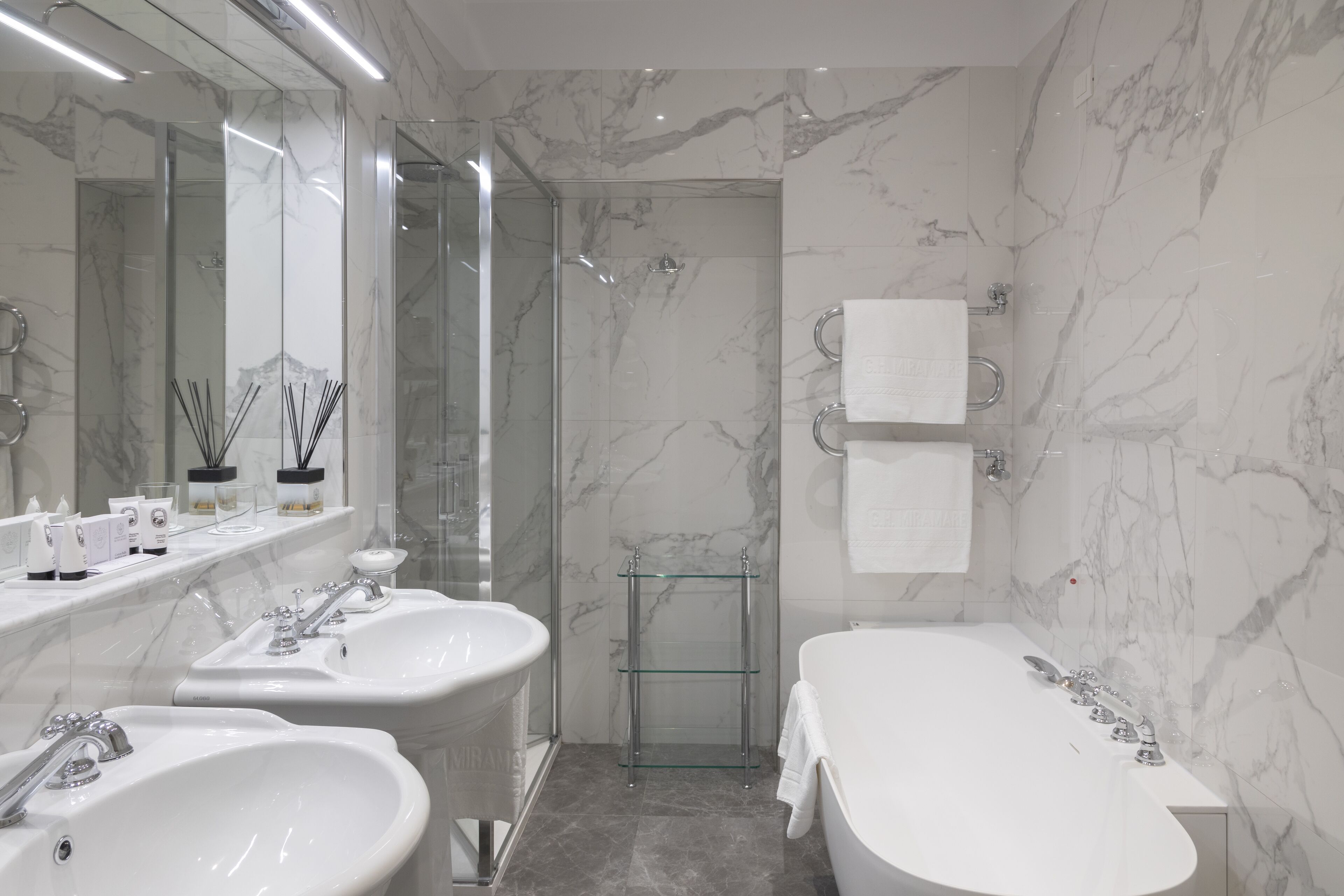 Une salle de bain moderne avec deux lavabos, une baignoire, une douche à l’italienne en verre et des murs en marbre.