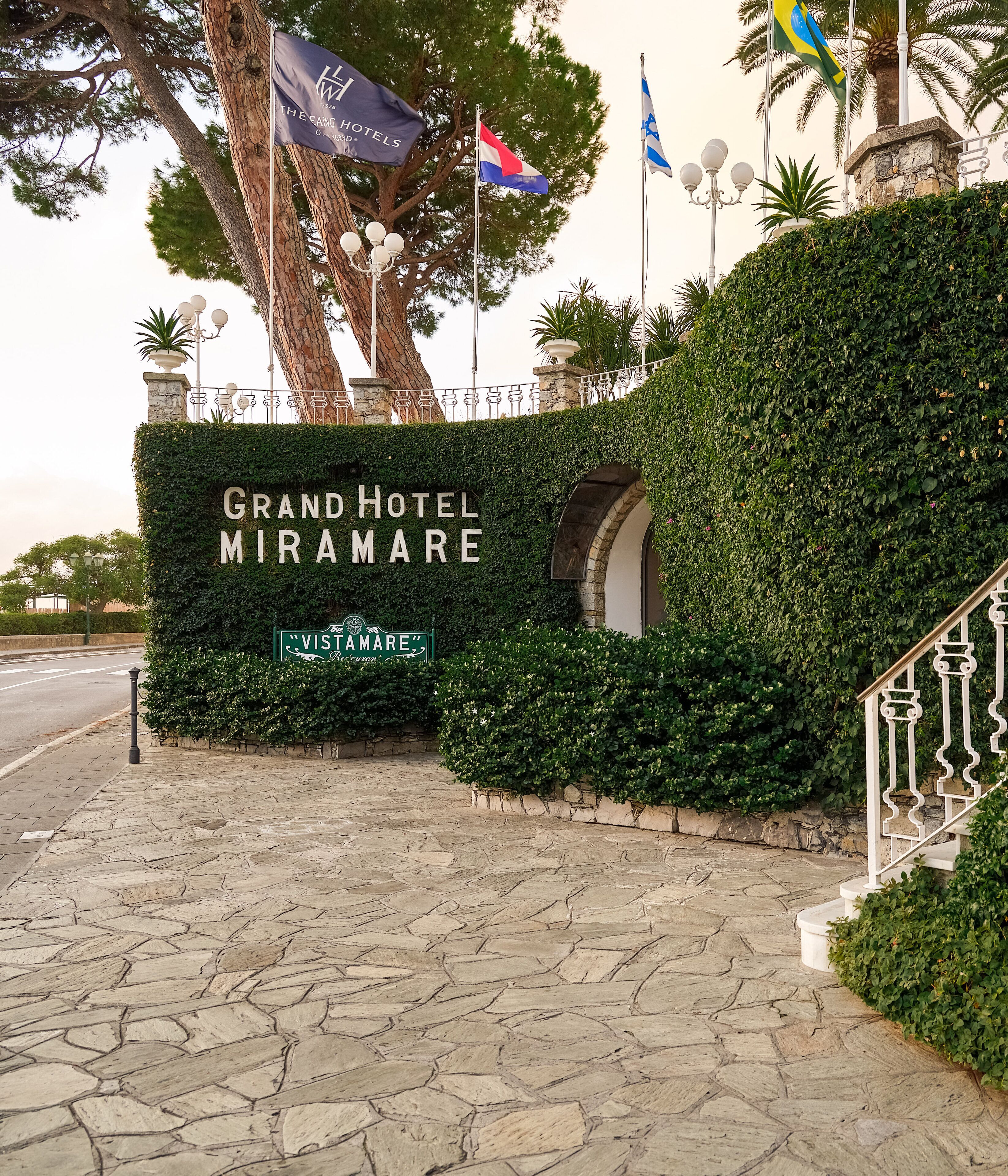 De ingang van Grand Hotel Miramare, met een stenen pad, een bord met de naam van het hotel en vlaggen.