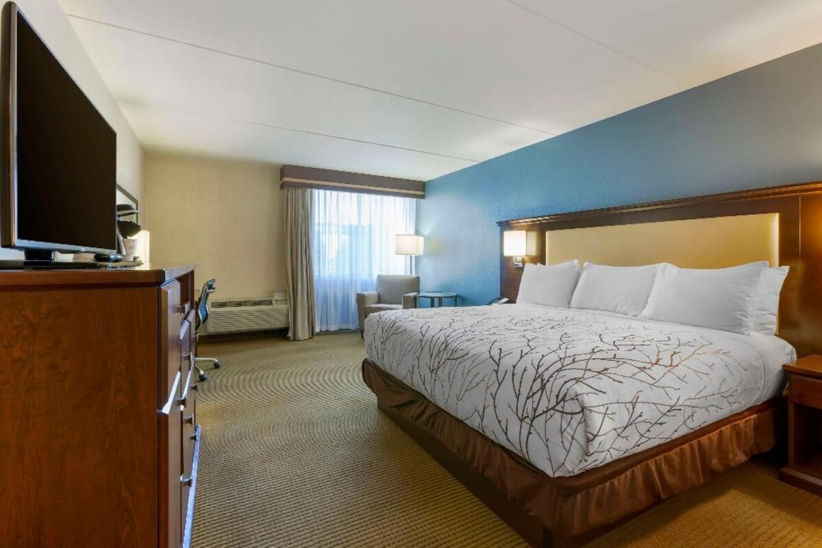 A hotel room with a large bed, a desk with a flat-screen TV, a lamp, and a window with curtains.