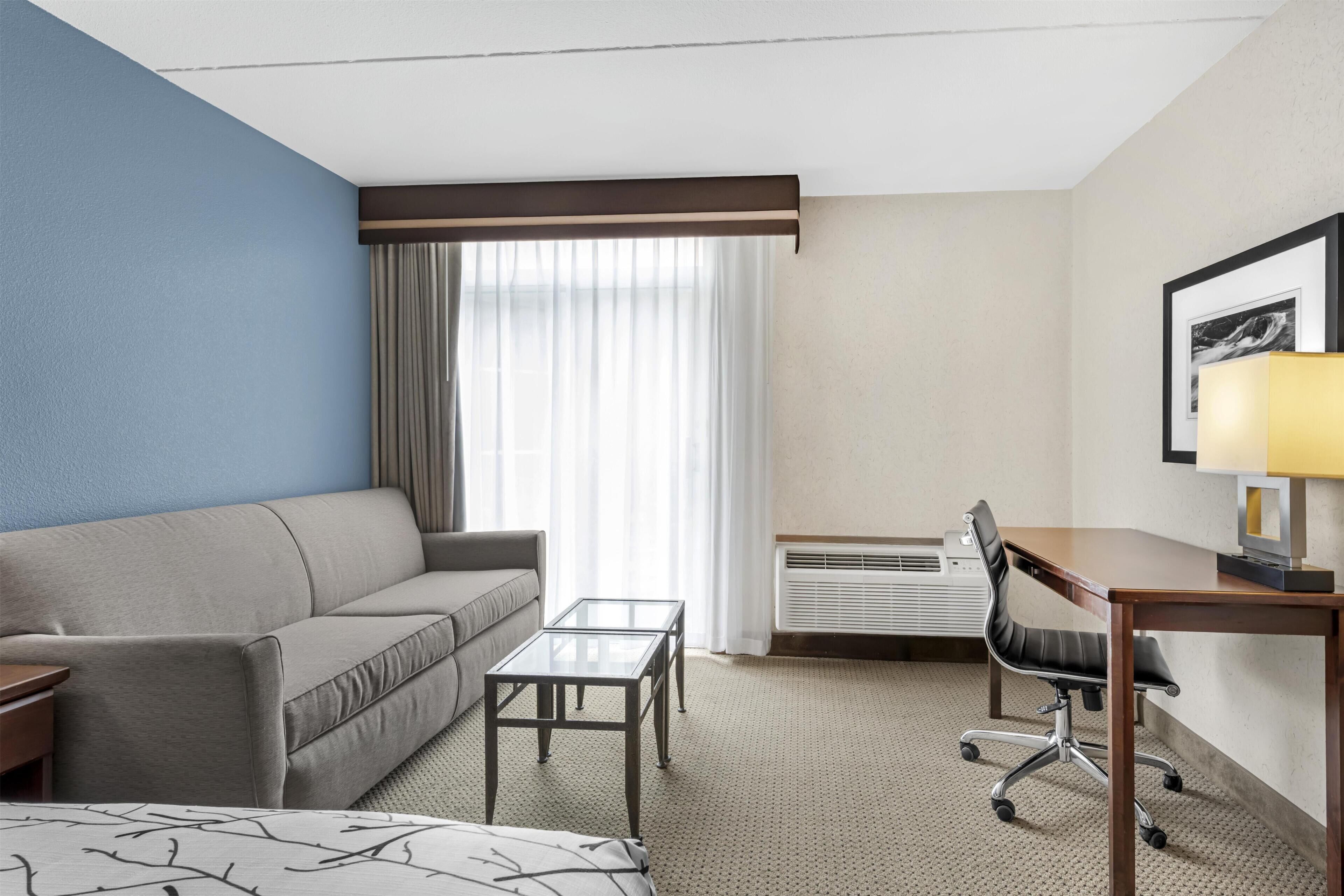A hotel room with a sofa, a small table, a desk with a chair, an air conditioning unit, and a window with curtains.