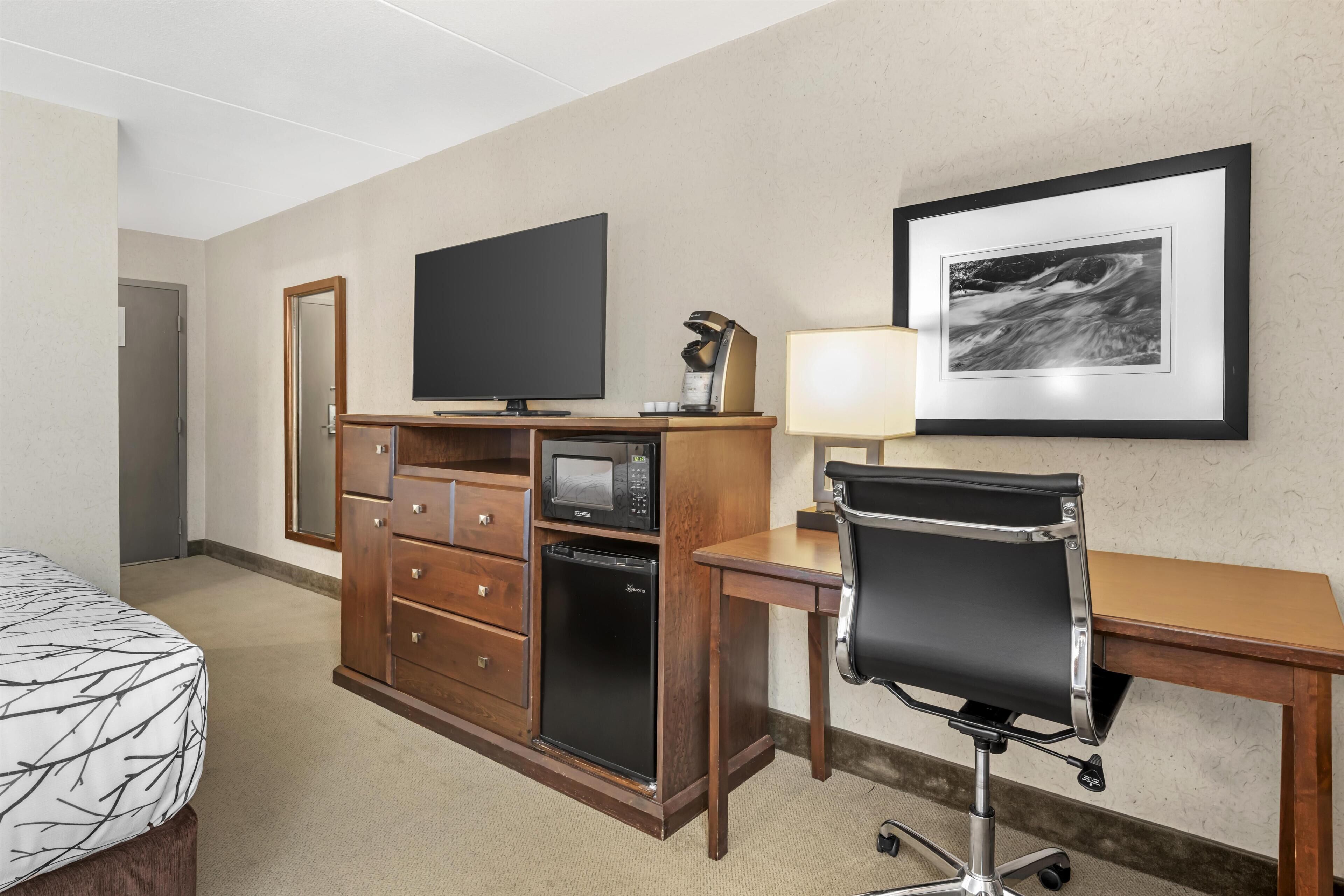 A hotel room with a bed, a desk with a chair, a television, a microwave, a refrigerator, a dresser, a lamp, and a framed picture on the wall.