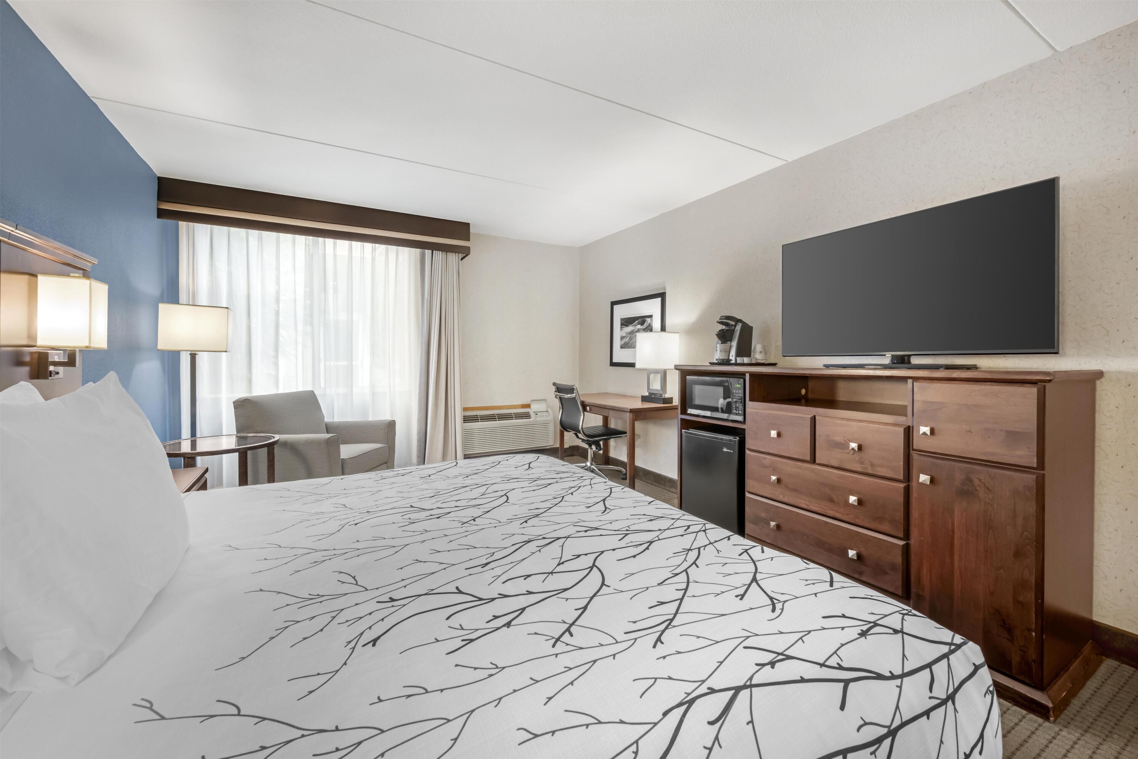 A hotel room with a large bed, a desk with a chair, a television, a wardrobe, and a window with curtains.
