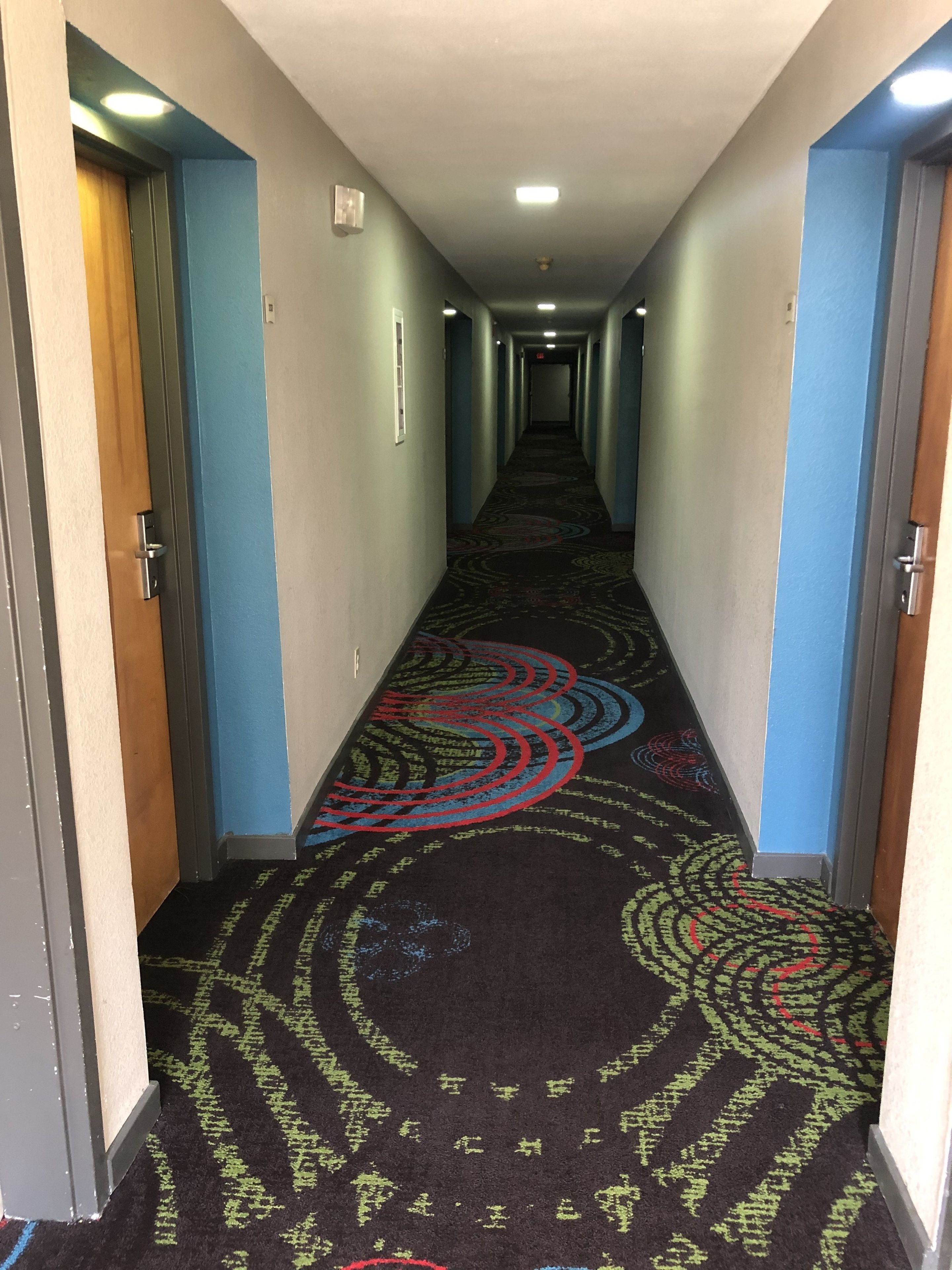 En hotellkorridor med ett mönstrat golv, blå väggar och en trädörr på vänster sida.