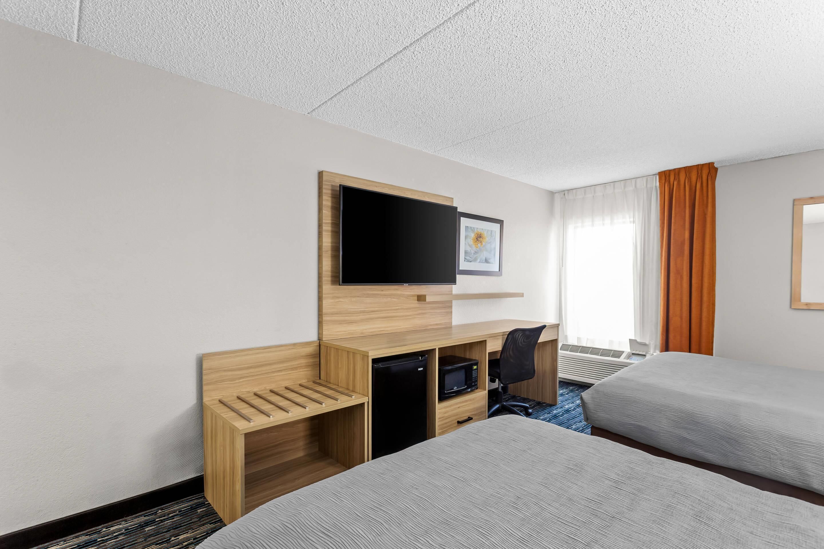 Ein Hotelzimmer mit zwei Betten, einem Schreibtisch mit Computer, einem Fernseher und einem Fenster mit Vorhängen.
