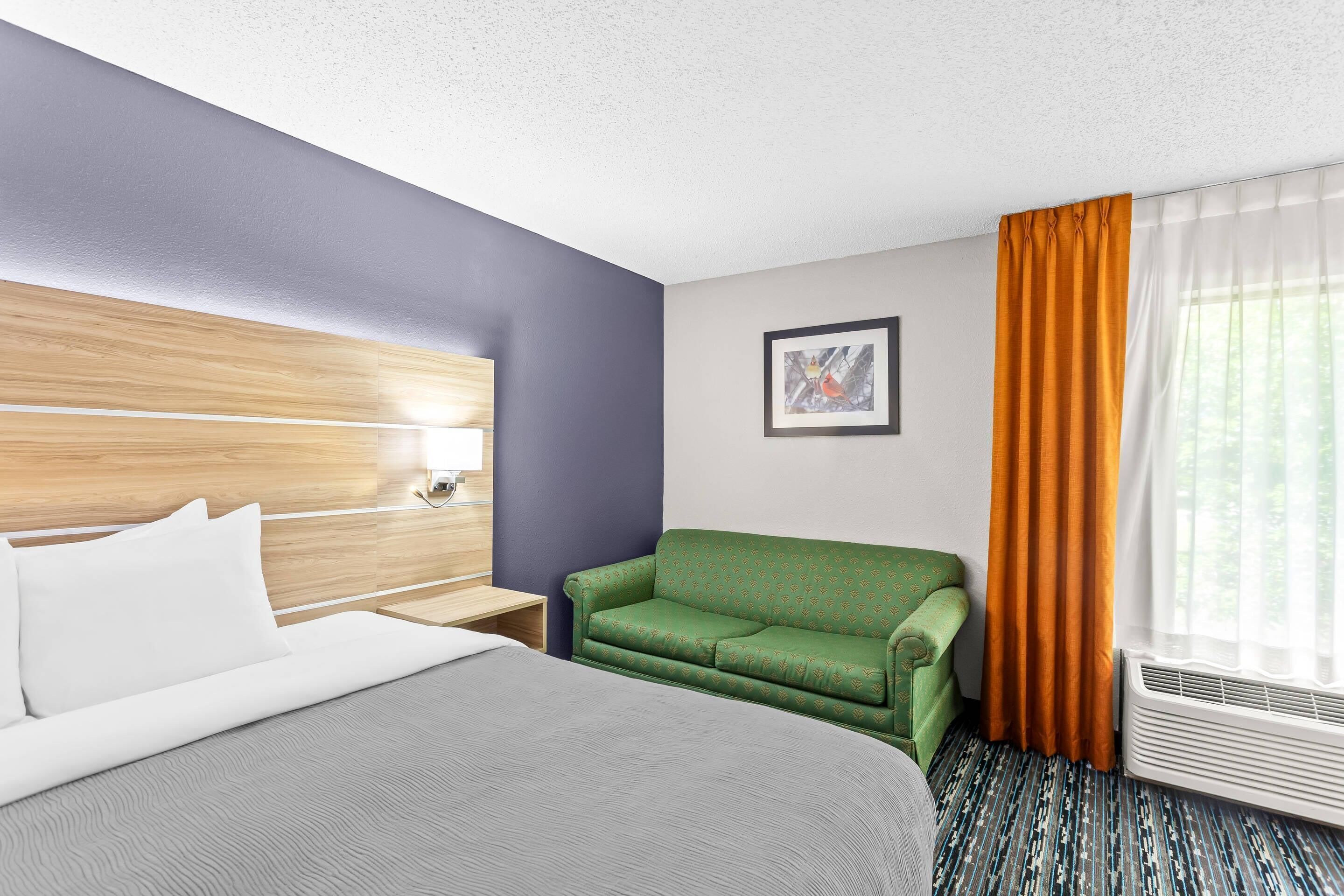 Ein Hotelzimmer mit einem Bett, einem grünen Sessel, einem Nachttisch, einem gerahmten Bild an der Wand und einem Fenster mit Vorhängen.