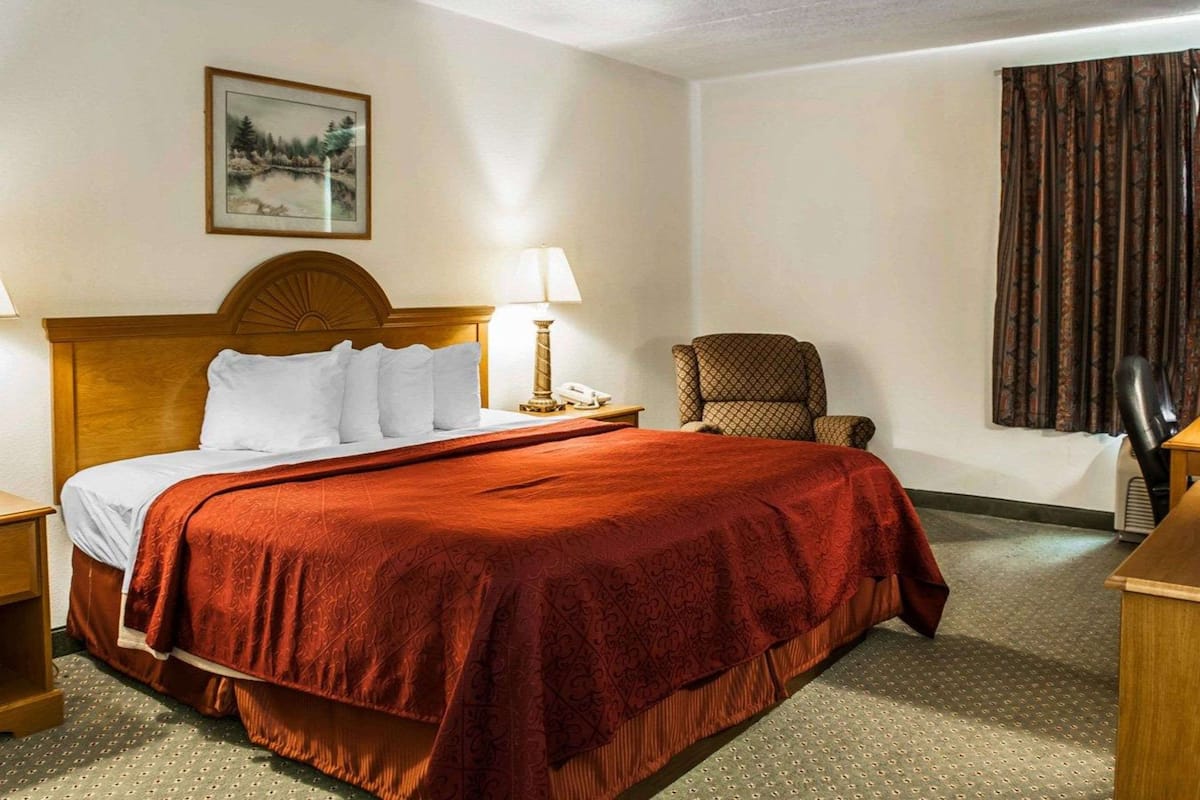 Quarto de hotel com cama grande, mesa de cabeceira, cadeira, abajur e um quadro na parede.