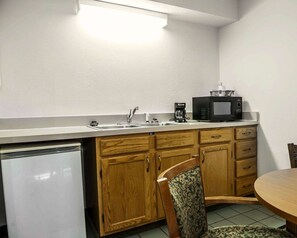 A compact kitchen with a refrigerator, sink, microwave, and coffee maker. There is a wooden cabinet with drawers and a chair with a patterned cushion.