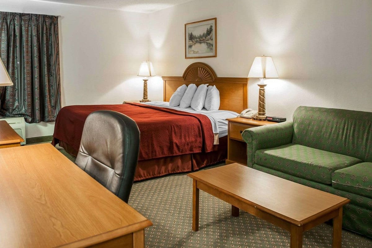 Quarto de hotel com uma cama, duas luminárias de cabeceira, um sofá verde, uma mesa de madeira e uma cadeira.