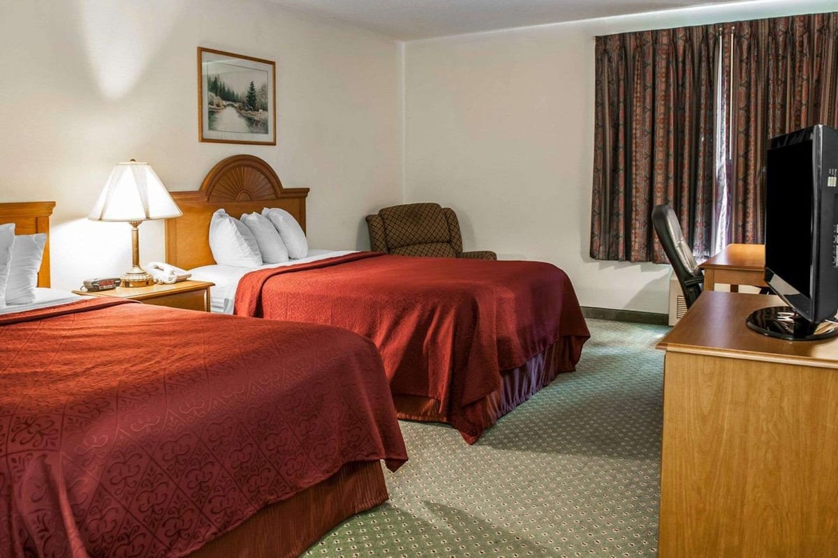 Quarto de hotel com duas camas, uma mesa com televisão, uma cadeira, um abajur e um quadro na parede.
