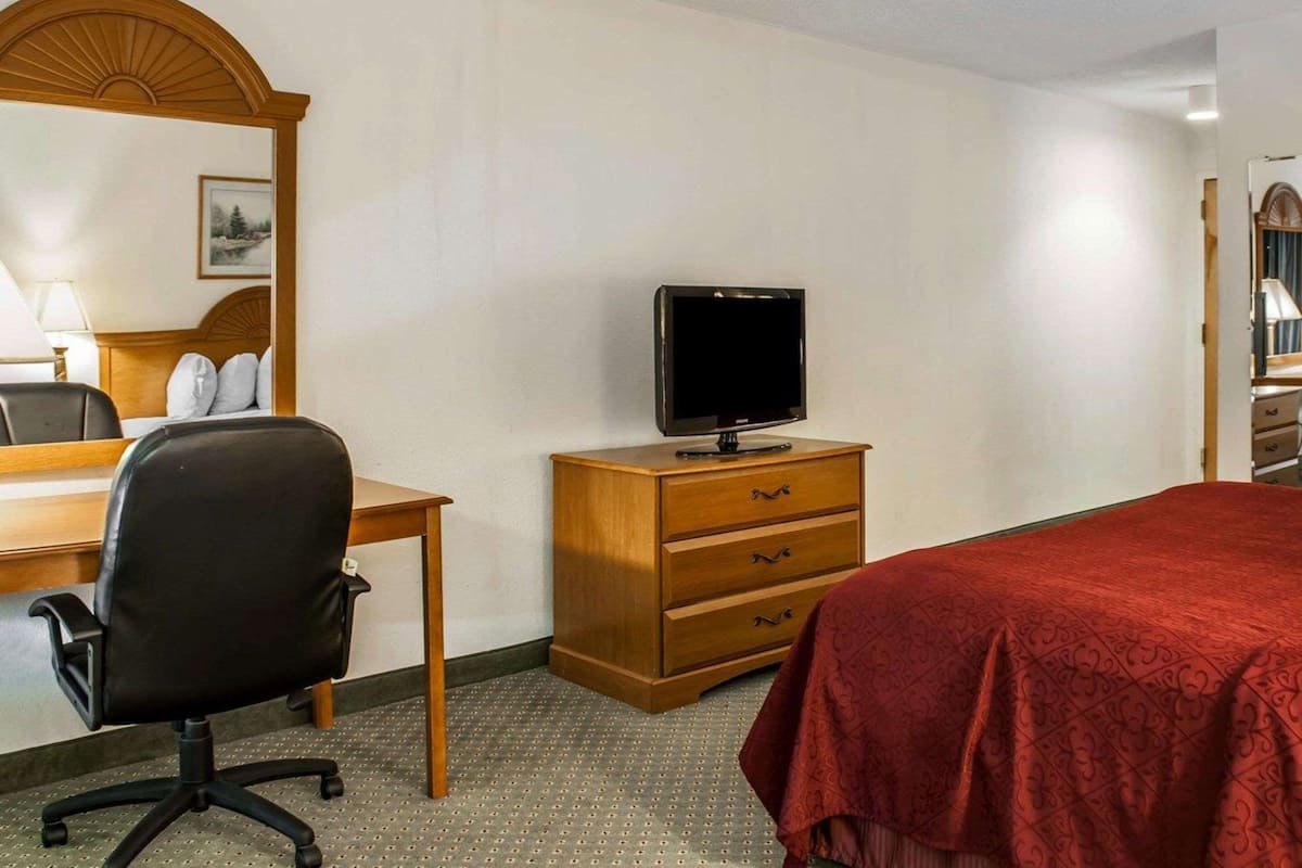 Quarto de hotel com cama, escrivaninha com cadeira, televisão em cómoda de madeira e espelho sobre a cómoda.