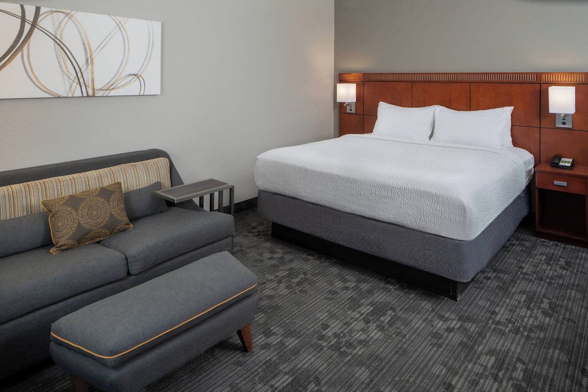 Une chambre d’hôtel avec un grand lit, un canapé, un pouf, une table de chevet et une lampe fixée au mur.