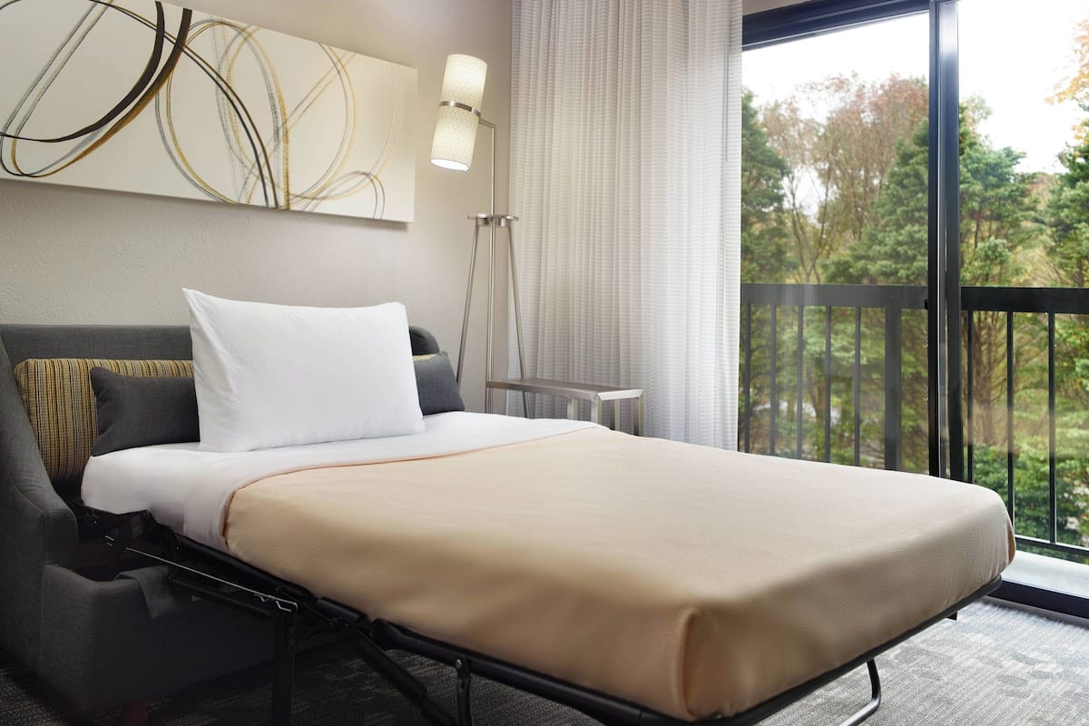 Une chambre d’hôtel avec un lit, une table de chevet, une lampe et une vue sur les arbres, visible à travers une grande fenêtre.