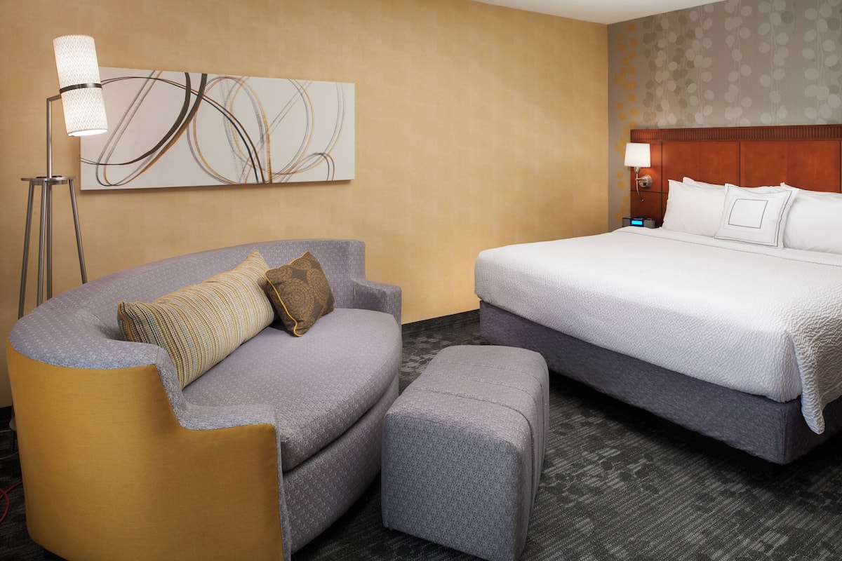 A hotel room with a curved sofa, a bed, a standing lamp, and a wall-mounted abstract artwork.