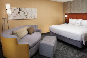 A hotel room with a curved sofa, a bed, a standing lamp, and a wall-mounted abstract artwork.