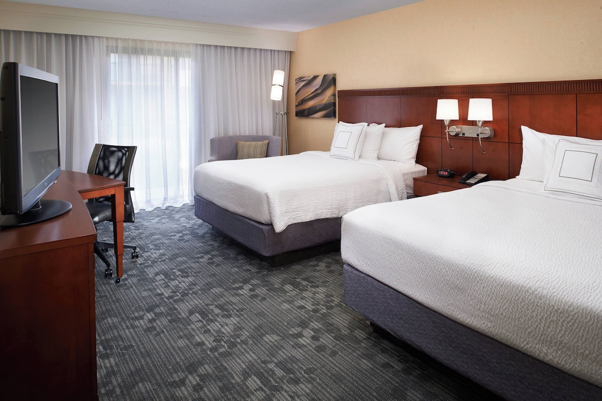 A hotel room with two beds, a desk with a television, a chair, and a nightstand with a phone and lamp.