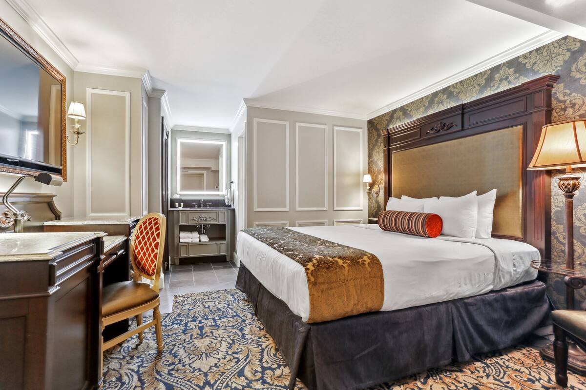 A hotel room with a large bed, a desk with a chair, a vanity with a mirror, and a patterned rug.