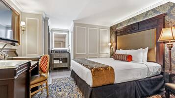 Een hotelkamer met een groot bed, een bureau met stoel, een kaptafel met spiegel en een kamerbreed tapijt met dessin.