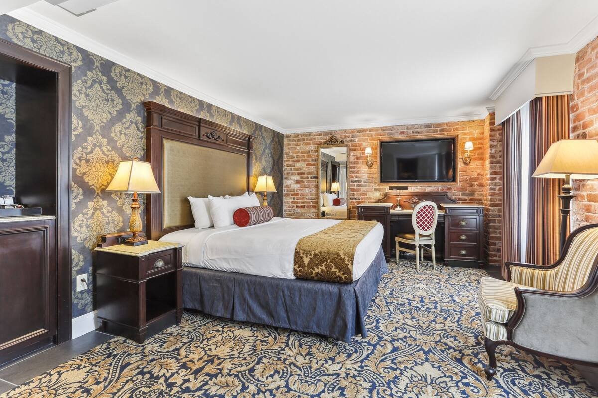 A hotel room with a large bed, two bedside tables with lamps, a desk with a chair, a TV mounted on a brick wall, and a patterned carpet.