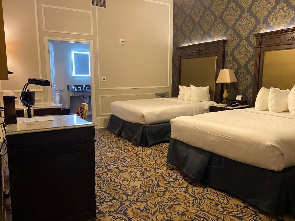A hotel room with two beds, a desk, a chair, and a bathroom visible through an open door.