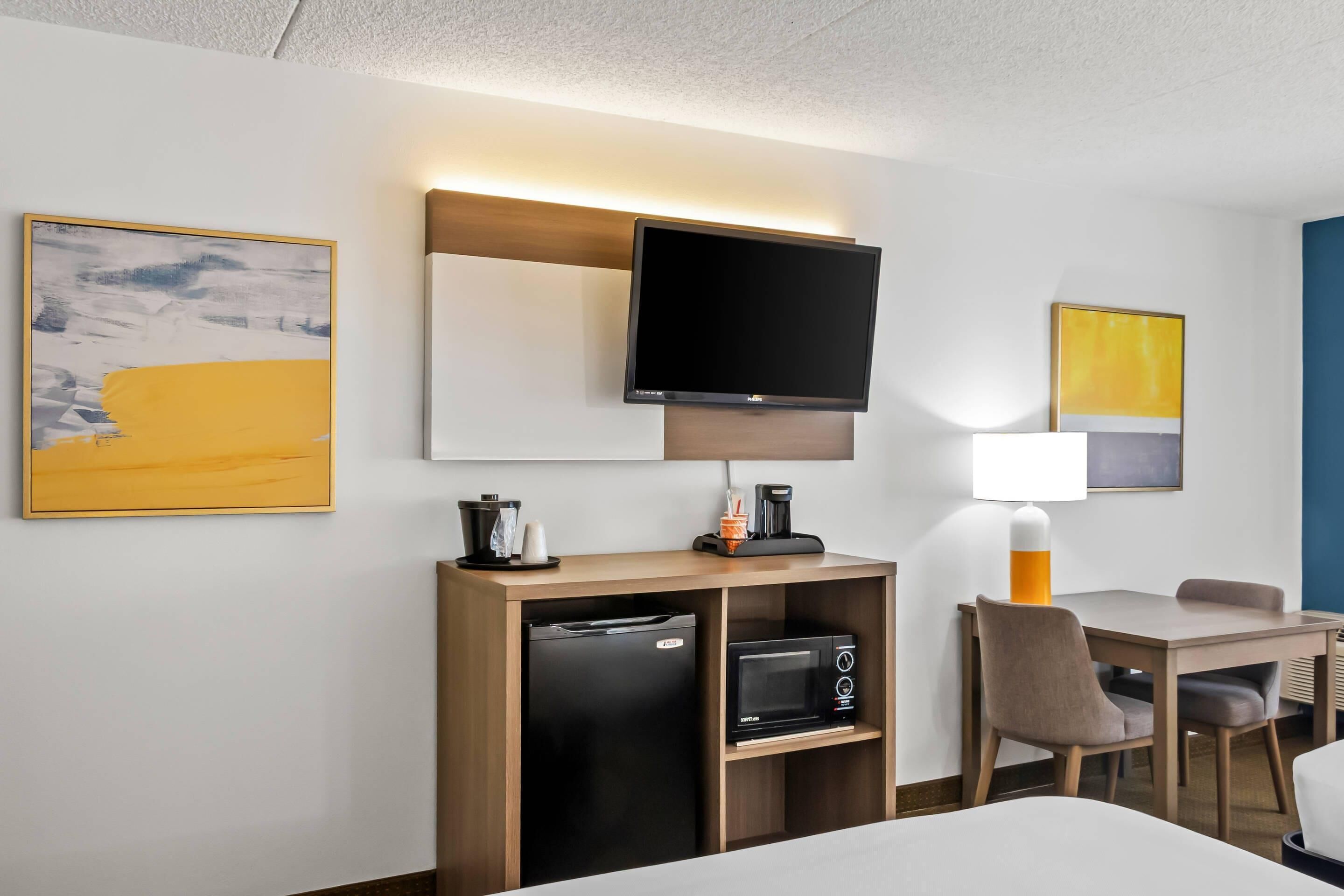 Ein Hotelzimmer mit Bett, Schreibtisch mit Kaffeemaschine, Mikrowelle, Fernseher und Kunstwerken an den Wänden.