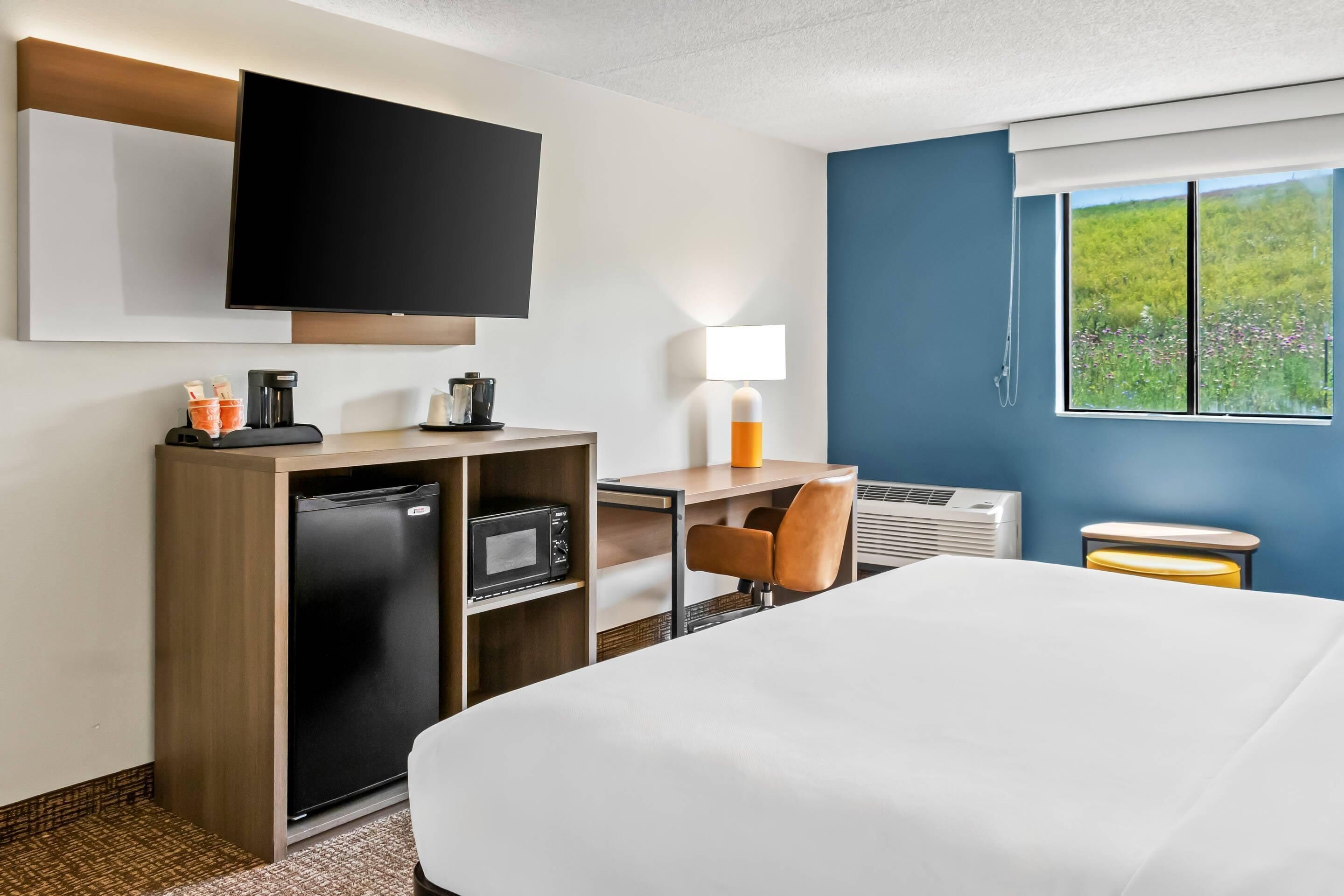 Habitación de hotel con cama, escritorio con computadora, televisor, refrigerador, cafetera y ventana con vista a áreas verdes.