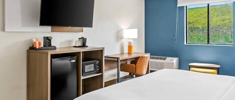 Habitación de hotel con cama, escritorio con computadora, televisor, refrigerador, cafetera y ventana con vista a áreas verdes.